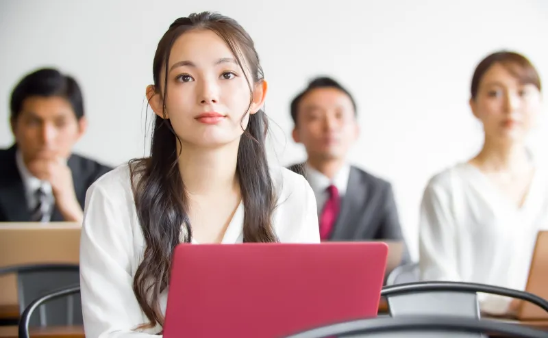 会議室でノートパソコンに向かう女性と、後ろに座る男性たち